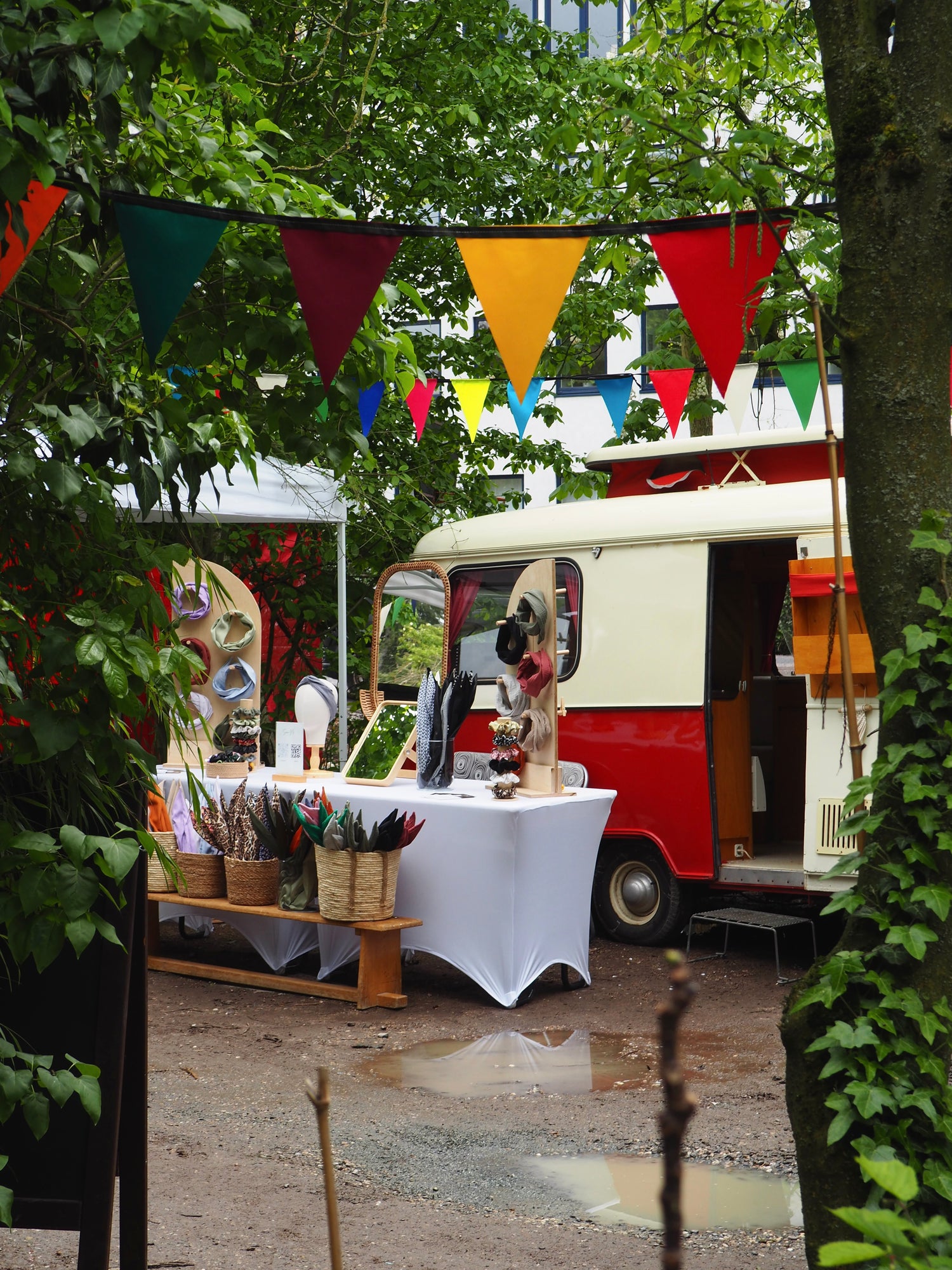 In einem sehr ausgiebig begrünten Innenhof ist ein liebevoll gestalteter Marktstand zu sehen, an dem Drahthaarbänder verkauft werden. Im Hintergrund ist ein nostalgischer Wohnwagen aus den 1960ern zu sehen. 