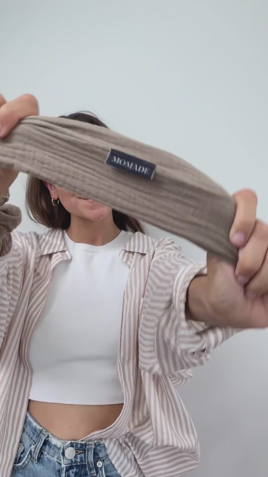Video laden: Vor einem weißen Hintergrund bindet sich eine Frau mit einem braunen Drahthaarband aus Musselinstoff eine Duttfrisur.